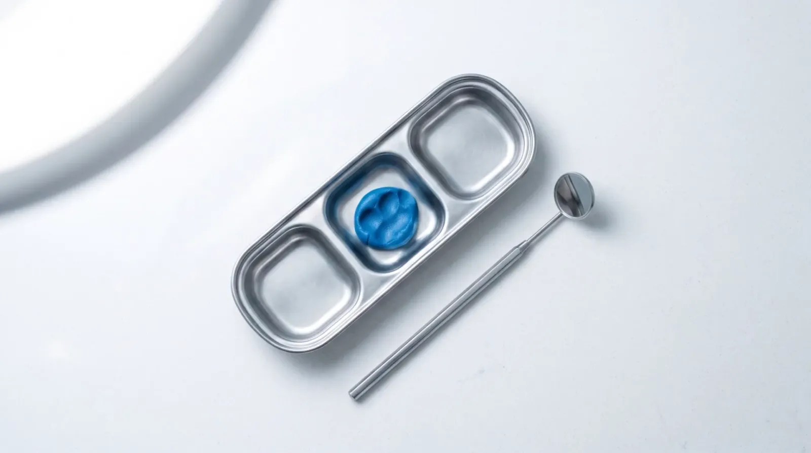 Illustrative overhead photograph of a stainless dental tray, set blue impression material and a mouth mirror.